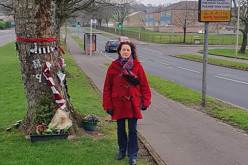Anne Hannigan standing next to memorial tree with Brediland Road in the background.
