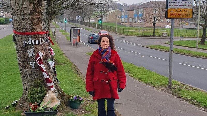 Anne Hannigan standing next to memorial tree with Brediland Road in the background.